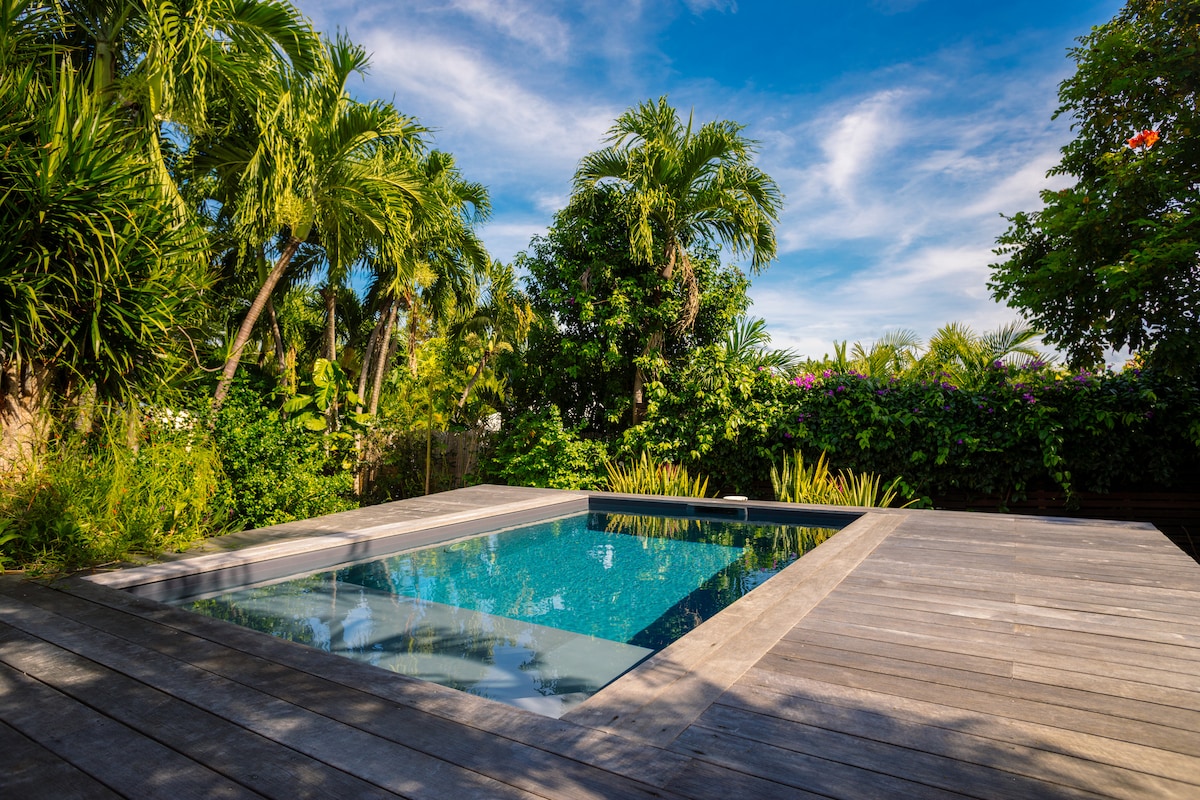 A serene pool area is surrounded by lush greenery and tropical plants. The crystal-clear water reflects the blue sky and is framed by a wooden deck, creating a peaceful outdoor space for relaxation.