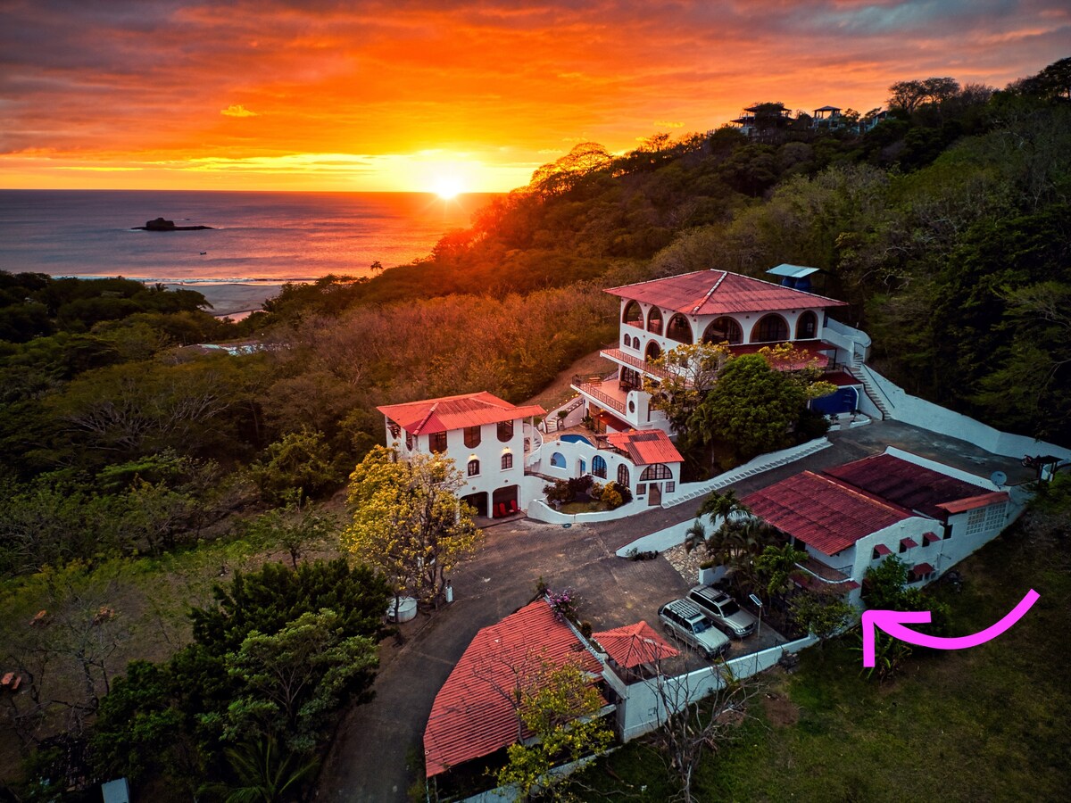 The image captures the ViYarte Villa overlooking the ocean at sunset, with vibrant colors filling the sky. The multi-level building features a red-tiled roof and is surrounded by lush greenery. A parking area is visible alongside the villa, with the beach in the background.