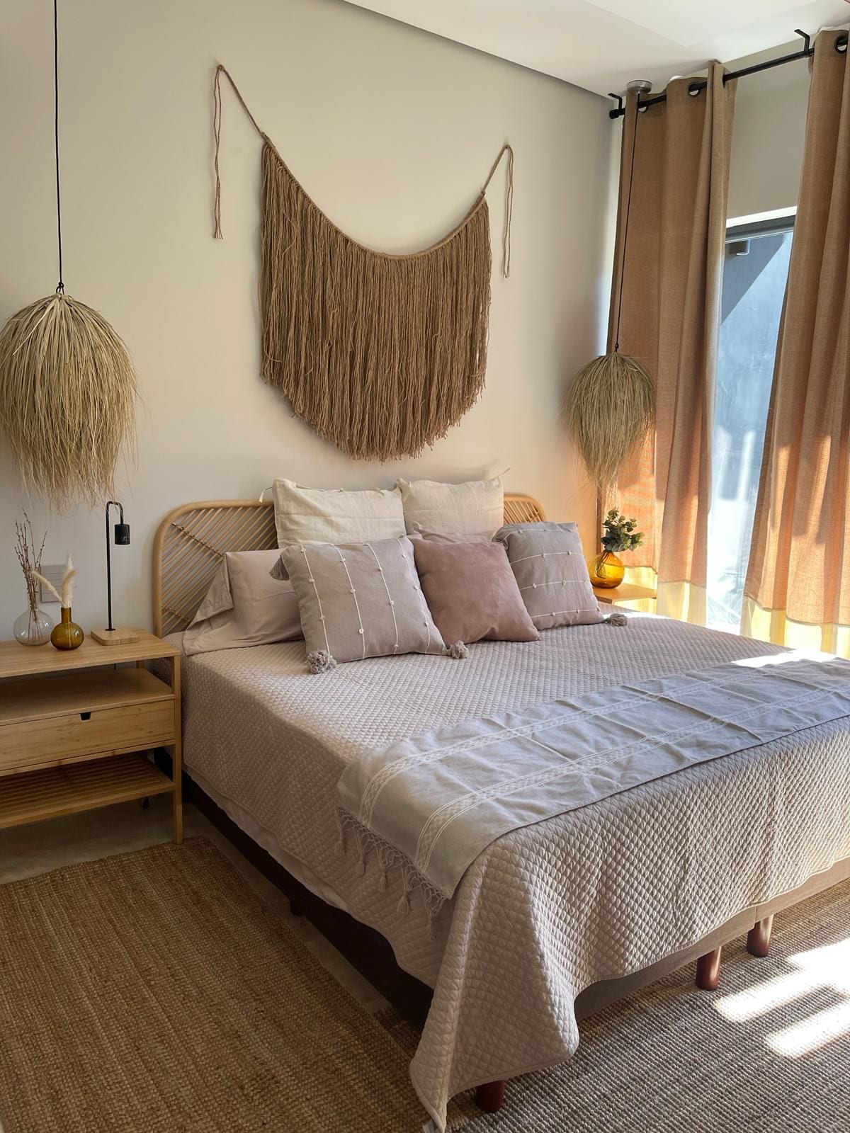 A cozy bedroom features a spacious bed adorned with various pillows in neutral tones. Soft textures are complemented by a woven wall hanging above the headboard, while natural light filters through curtains. A bedside table and decorative plants add subtle charm to the serene environment.