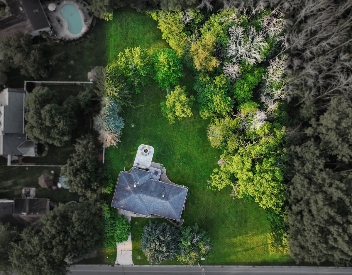 An aerial view captures the ranch-style house surrounded by lush greenery and trees. The expansive yard features a blend of manicured grass and natural wooded areas, providing ample outdoor space for gatherings and privacy.