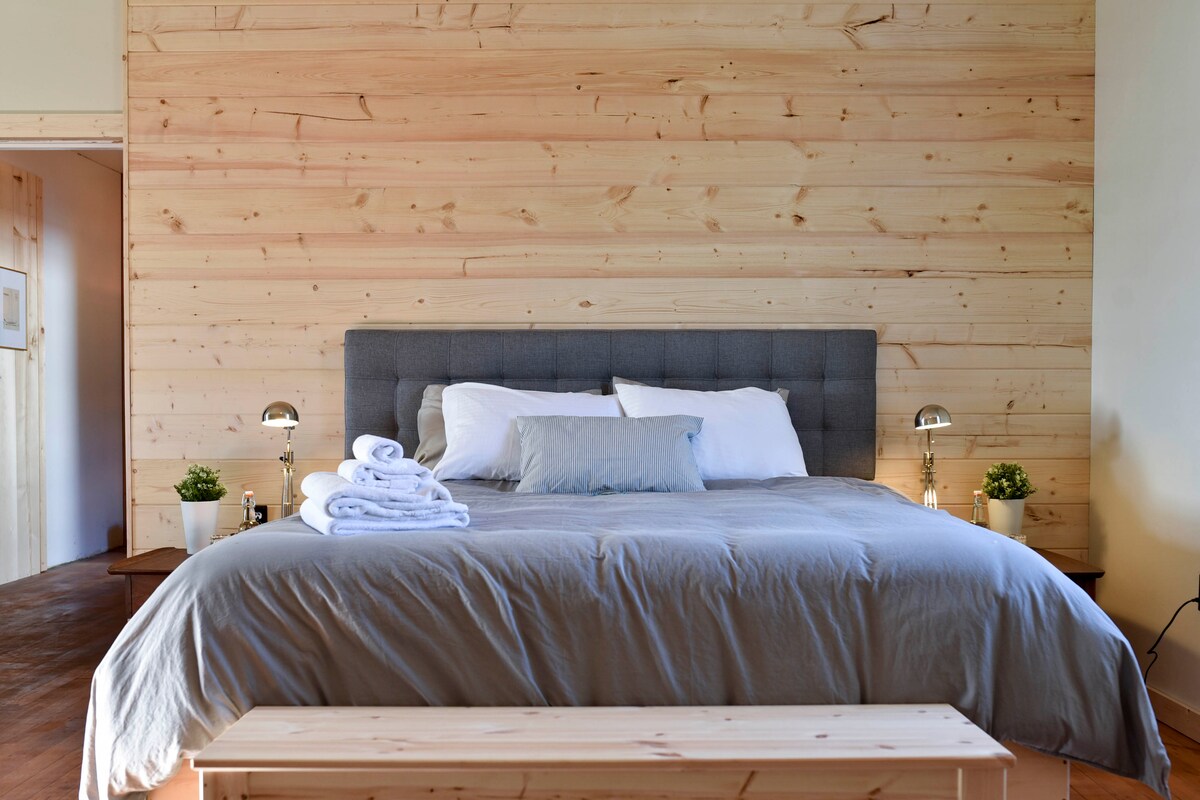 A cozy bedroom features a spacious bed adorned with neutral bedding and decorative pillows. Soft lighting from bedside lamps illuminates the wooden wall behind the bed, complemented by small potted plants on either side. A wooden bench is positioned at the foot of the bed.