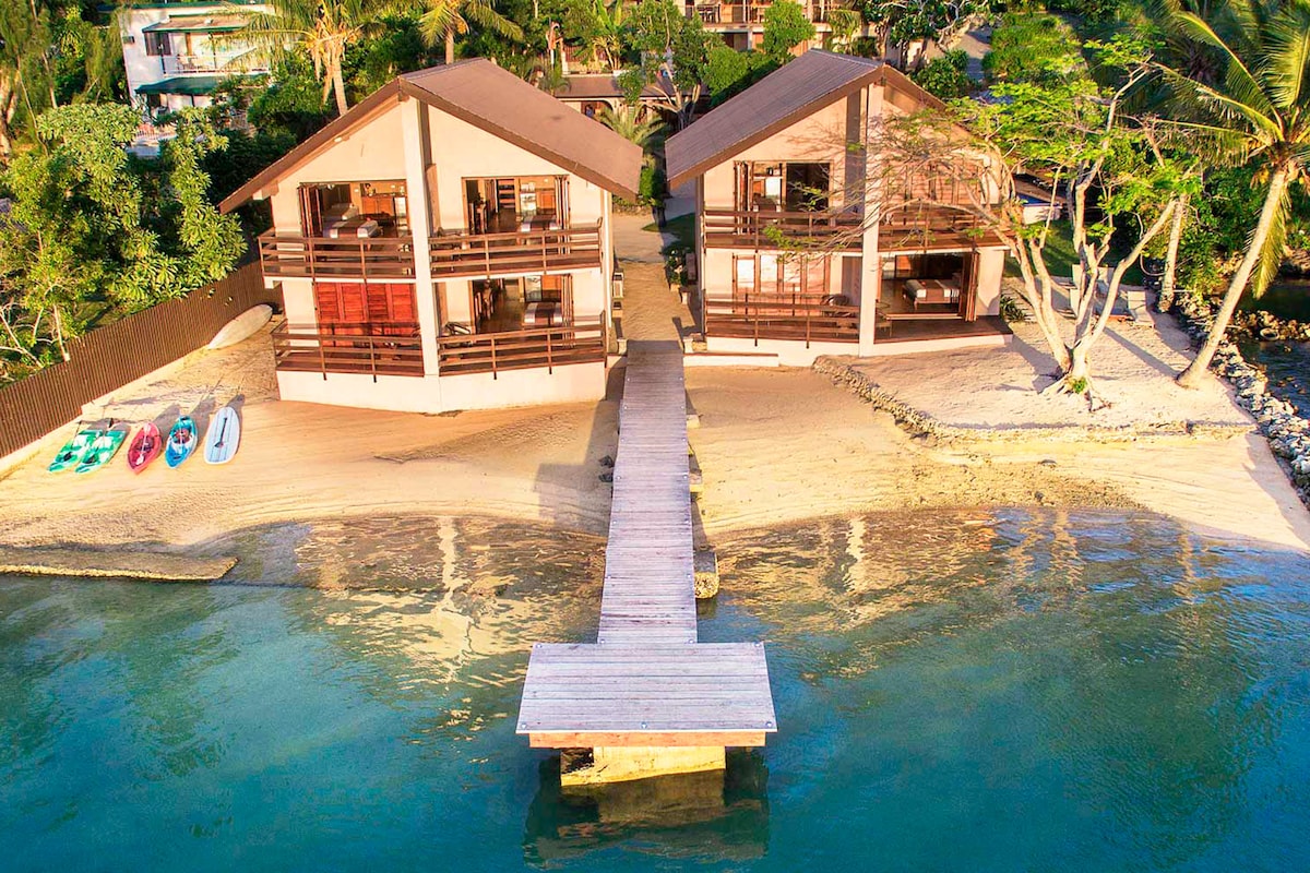 Two contemporary boutique hotel buildings are seen from above, set on a sandy beach. A wooden walkway extends from the beach to the water, while kayaks and stand-up paddleboards rest nearby. Lush greenery surrounds the area, enhancing the serene coastal environment.