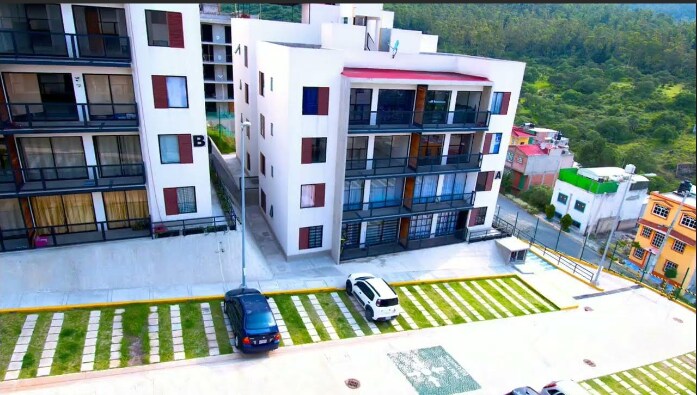 A modern building complex is depicted, featuring multiple levels with balconies. The ground level includes a paved parking area and neatly arranged green grass. The surrounding landscape showcases greenery, indicating a natural environment. The architecture presents a clean and contemporary aesthetic.