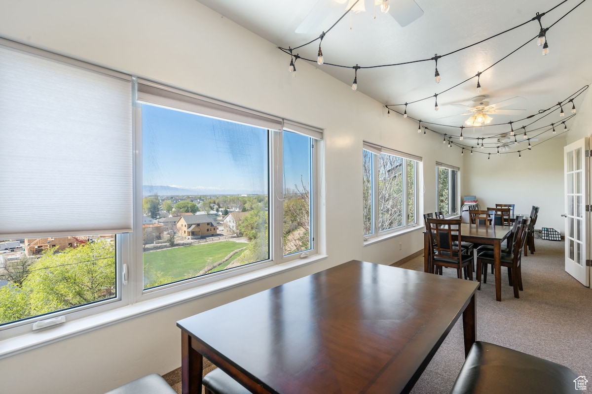 A bright dining area is filled with natural light from large windows, offering views of the surrounding landscape. A spacious wooden table is centered in the room, accompanied by several chairs. Decorative string lights hang overhead, creating a relaxed ambiance.