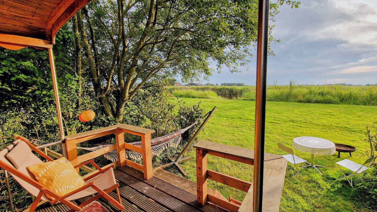 A wooden deck area is shown, featuring a hammock and a lounge chair. A view of the expansive green field and trees is visible, with two white chairs and a small table set in the grass. Natural light highlights the inviting outdoor seating space.