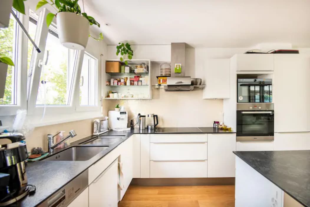 A modern open kitchen is featured, equipped with stainless steel appliances, including two ovens and a dishwasher. Ample counter space accommodates various kitchen gadgets. Natural light fills the area through multiple windows, and shelves display jars and cooking essentials along the wall.