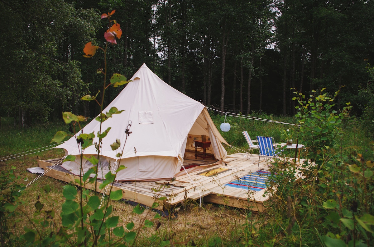 A glamping tent is positioned on a wooden platform, surrounded by lush greenery. Two outdoor chairs are visible next to the tent, and a lantern hangs above the entrance, providing a cozy touch. The peaceful forest backdrop enhances the natural setting.