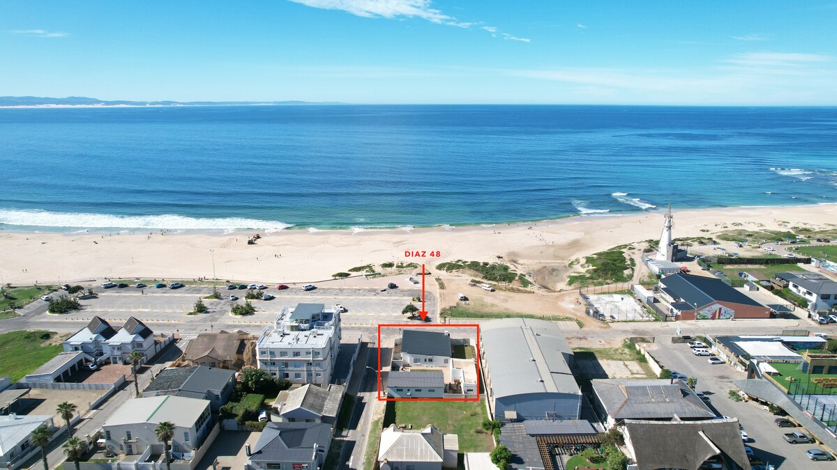 An aerial view captures the property set against a backdrop of the ocean, with sandy beaches stretching along the coastline. The clear blue water contrasts with the rooftops of nearby homes, and the lighthouse stands prominently in the distance.