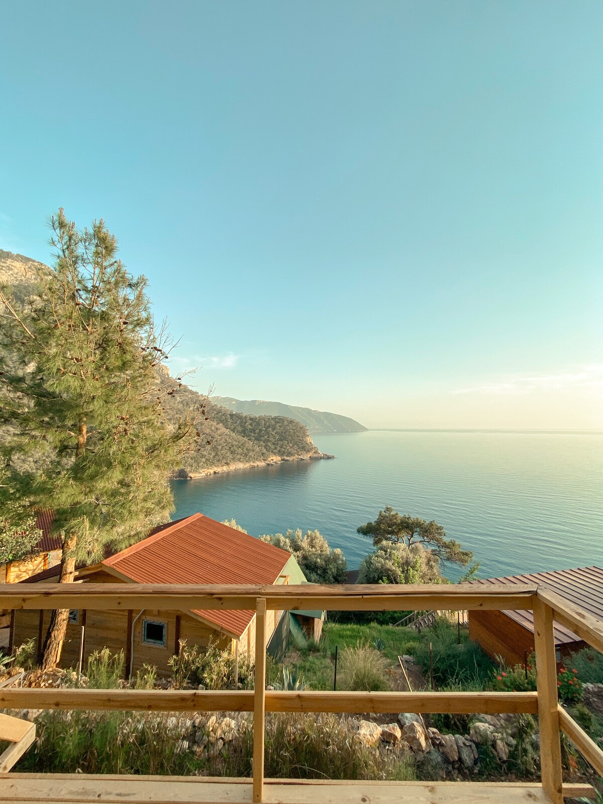 A serene view of the coastline is displayed, featuring a calm blue sea bordered by green hills. The foreground showcases wooden structures with red roofs, surrounded by lush vegetation. The sky is clear with soft colors, enhancing the tranquil atmosphere of the surroundings.
