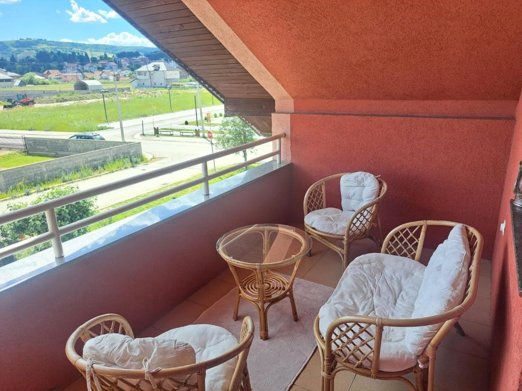 A cozy balcony area is highlighted by a small glass table surrounded by four rattan chairs with soft white cushions. The terracotta walls provide warmth, while a view of the green fields and distant buildings is visible beyond the railing.