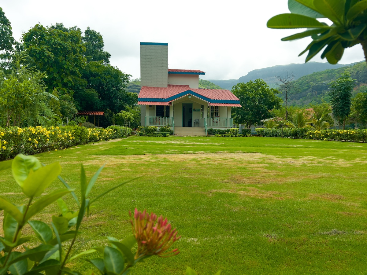 The front view of the villa showcases a modern two-story structure with a red-tiled roof, framed by lush greenery and colorful flowers. A spacious lawn leads to the entrance, complemented by gentle hills in the background under a cloudy sky.