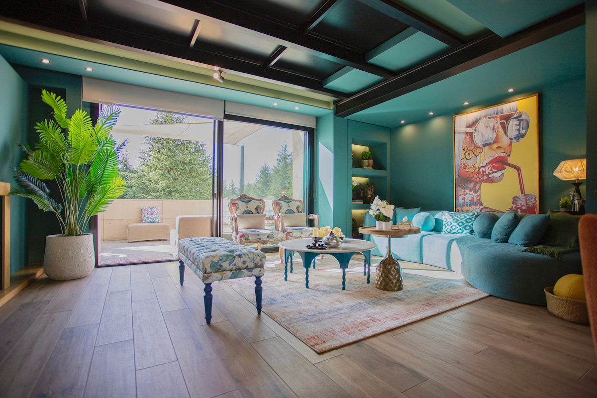 A spacious living area features teal walls and stylish furniture, including a large, rounded sofa and a patterned bench. Natural light streams in through glass doors that open to a terrace, while a vibrant piece of wall art adds a pop of color.