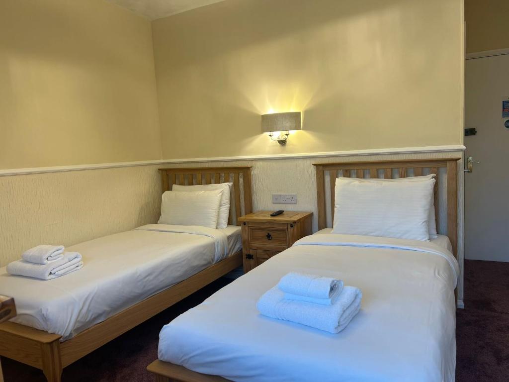 A well-lit room features two single beds with white linens, accompanied by neatly folded towels. A wooden nightstand is positioned between the beds, and gentle wall lighting adds a soft glow to the inviting atmosphere.