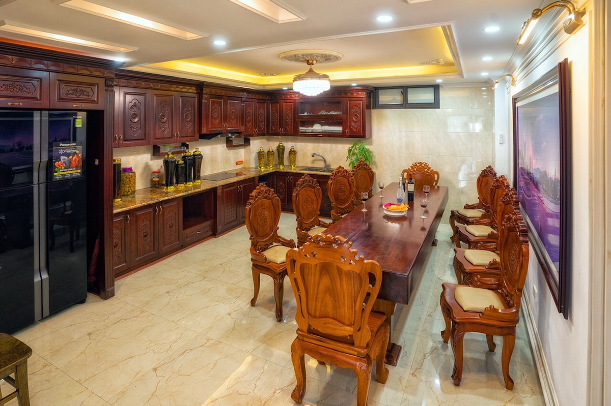 A spacious kitchen and dining area features dark wooden cabinetry and a large dining table surrounded by intricately carved chairs. The glossy marble floor reflects warm lighting from a chandelier above, and kitchen appliances are neatly arranged along the counters.