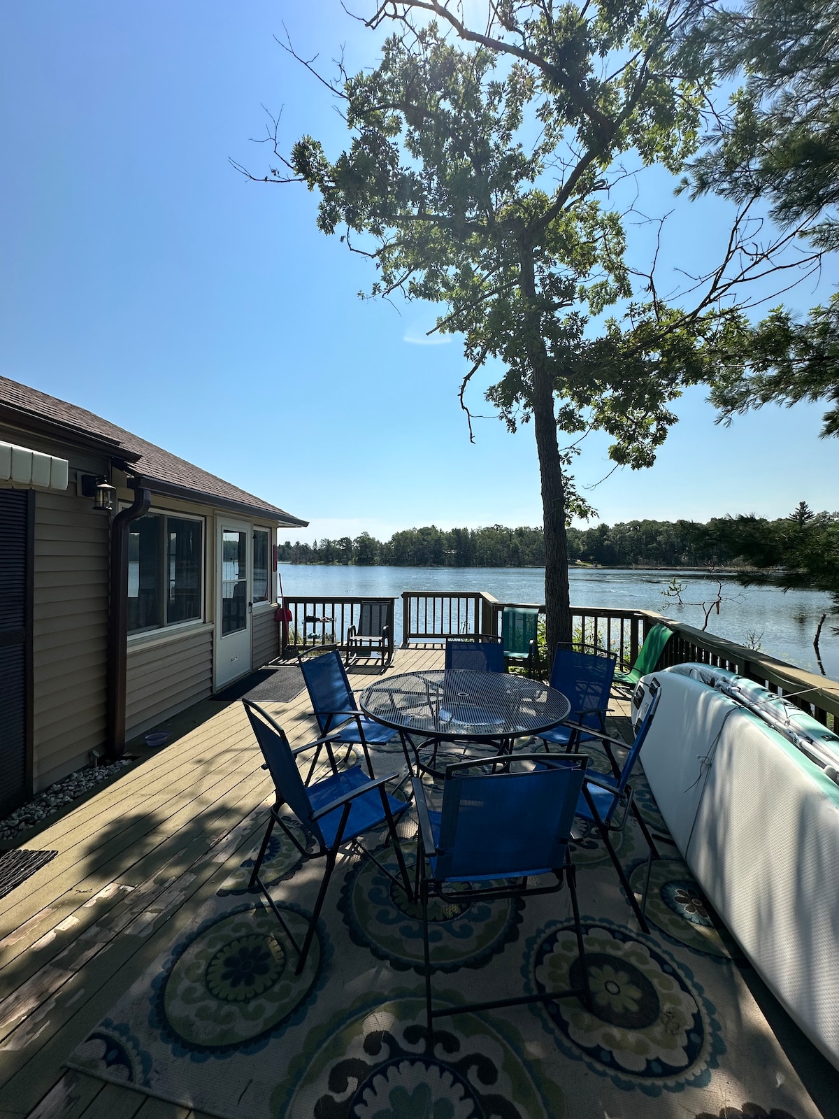 A spacious deck overlooks the tranquil waters of Long Lake. The area is furnished with a round table surrounded by six blue chairs, inviting guests to enjoy outdoor dining. A large tree provides shade, enhancing the serene lake view under a clear blue sky.