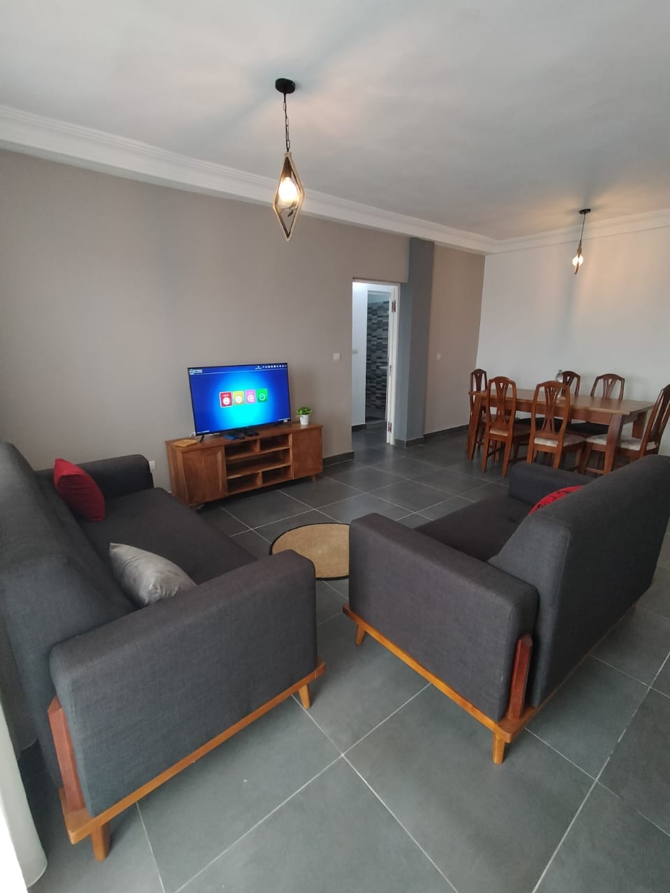 A cozy living area features two dark gray sofas positioned across from a wooden TV stand, where a television is displayed. A round area rug sits on the tiled floor, complementing the minimalist decor. A dining table with several chairs is visible in the background.