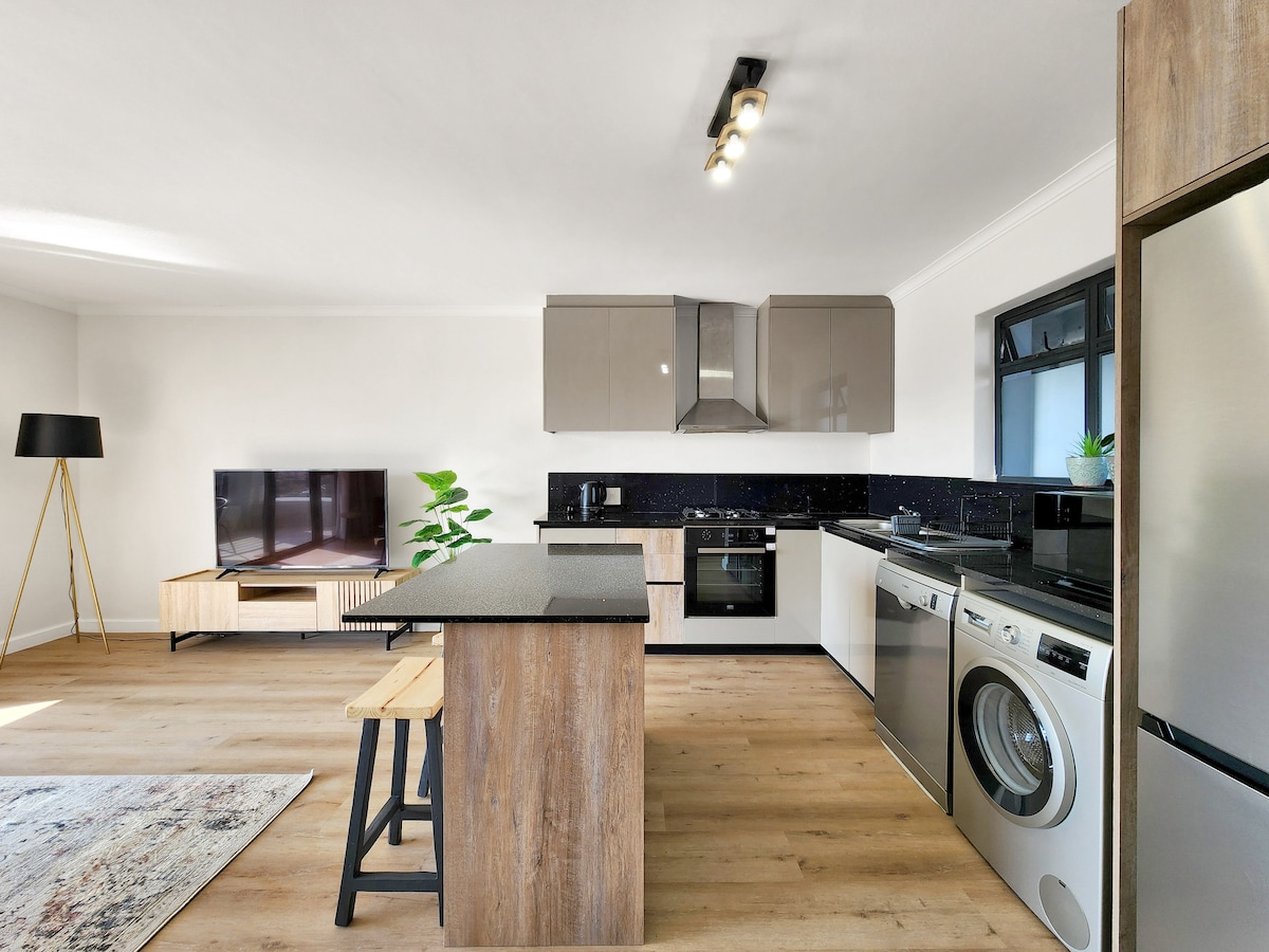 A modern kitchen is showcased, featuring contemporary cabinets and sleek appliances. An island counter with seating provides a casual dining option, while a flat-screen TV is positioned in the living area. Natural light fills the space, enhancing the clean, inviting atmosphere.