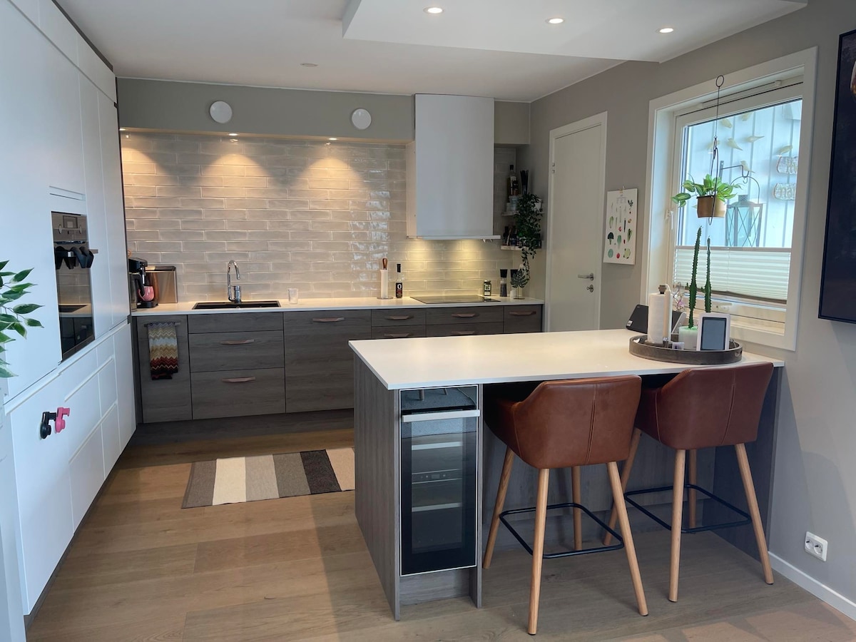 A modern kitchen is showcased, featuring a central island with two brown upholstered chairs. Light-colored cabinetry and a textured backsplash enhance the space. Under-cabinet lighting illuminates the countertops, while natural light enters through a nearby window, creating a bright and functional environment.