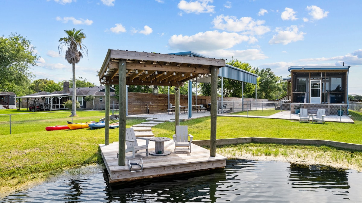 A waterfront structure features a shaded seating area on a wooden platform, surrounded by grass and calm water. Colorful kayaks are visible on the edge of the lawn, while the background includes a fenced area and several seating options under a blue sky.