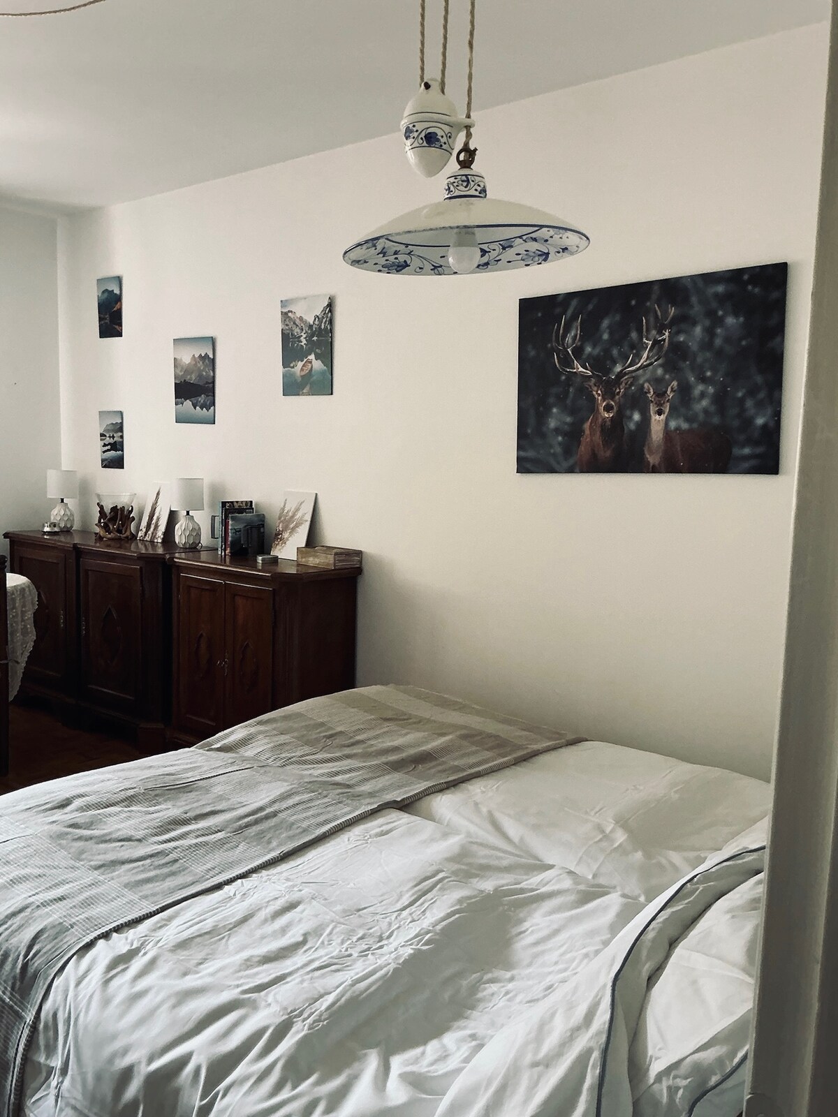 A well-appointed bedroom features a spacious bed with light linens, complemented by a tasteful pendant light. Artwork of serene landscapes adorns the walls, while a wooden cabinet provides additional storage, enhancing the cozy ambiance of the room.