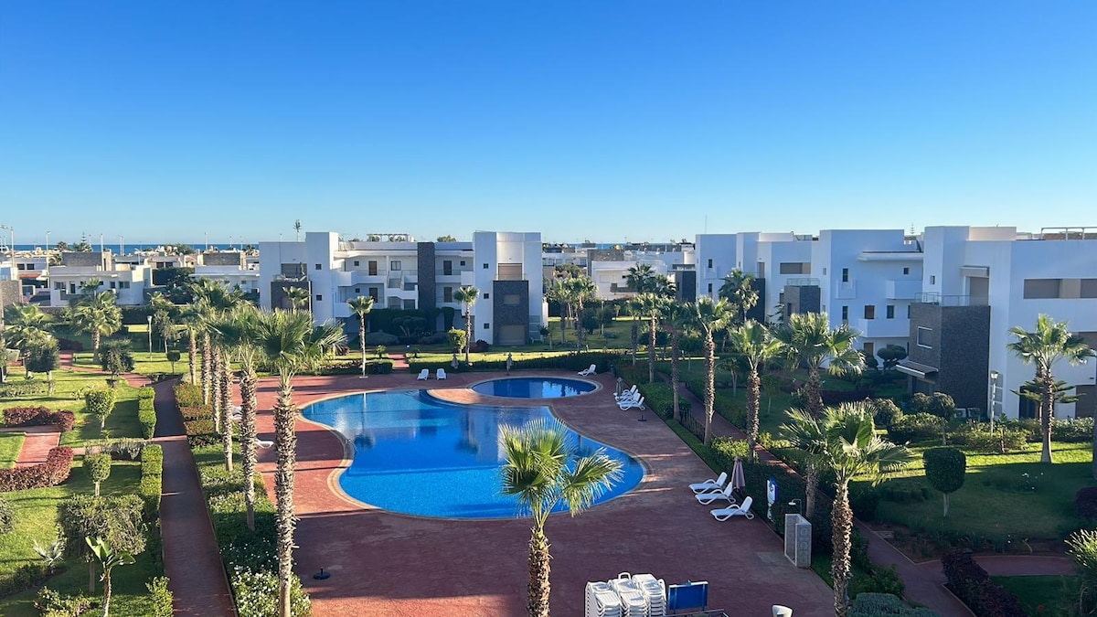 A serene view captures a well-maintained community featuring a large swimming pool surrounded by lush palm trees. White buildings create a modern environment, while vibrant greenery lines the walkways, contributing to a tranquil atmosphere.