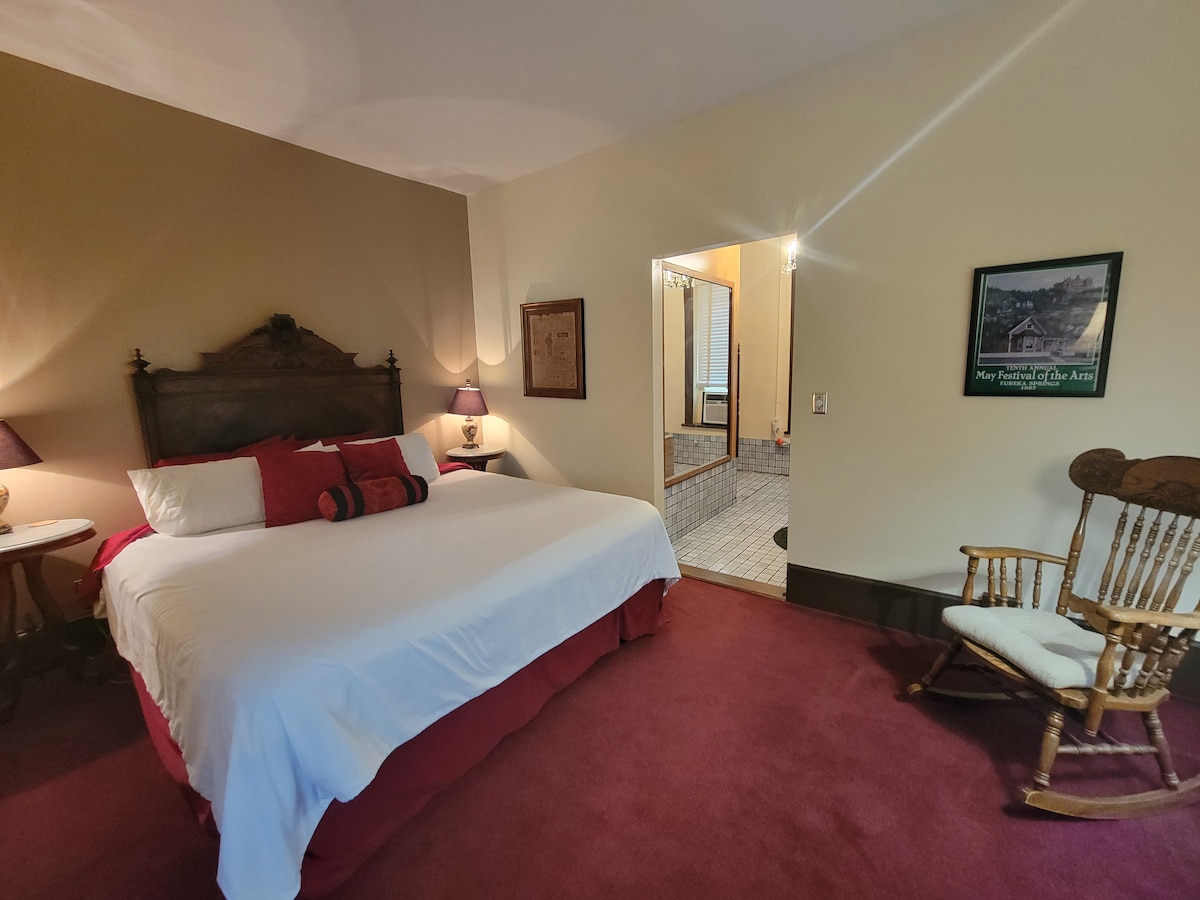 A spacious bedroom is presented, featuring a plush king-sized bed dressed in white linens with red accents. Soft lighting is provided by bedside lamps. A rocking chair is positioned nearby, while an open doorway leads to a bathroom area with tiled flooring.