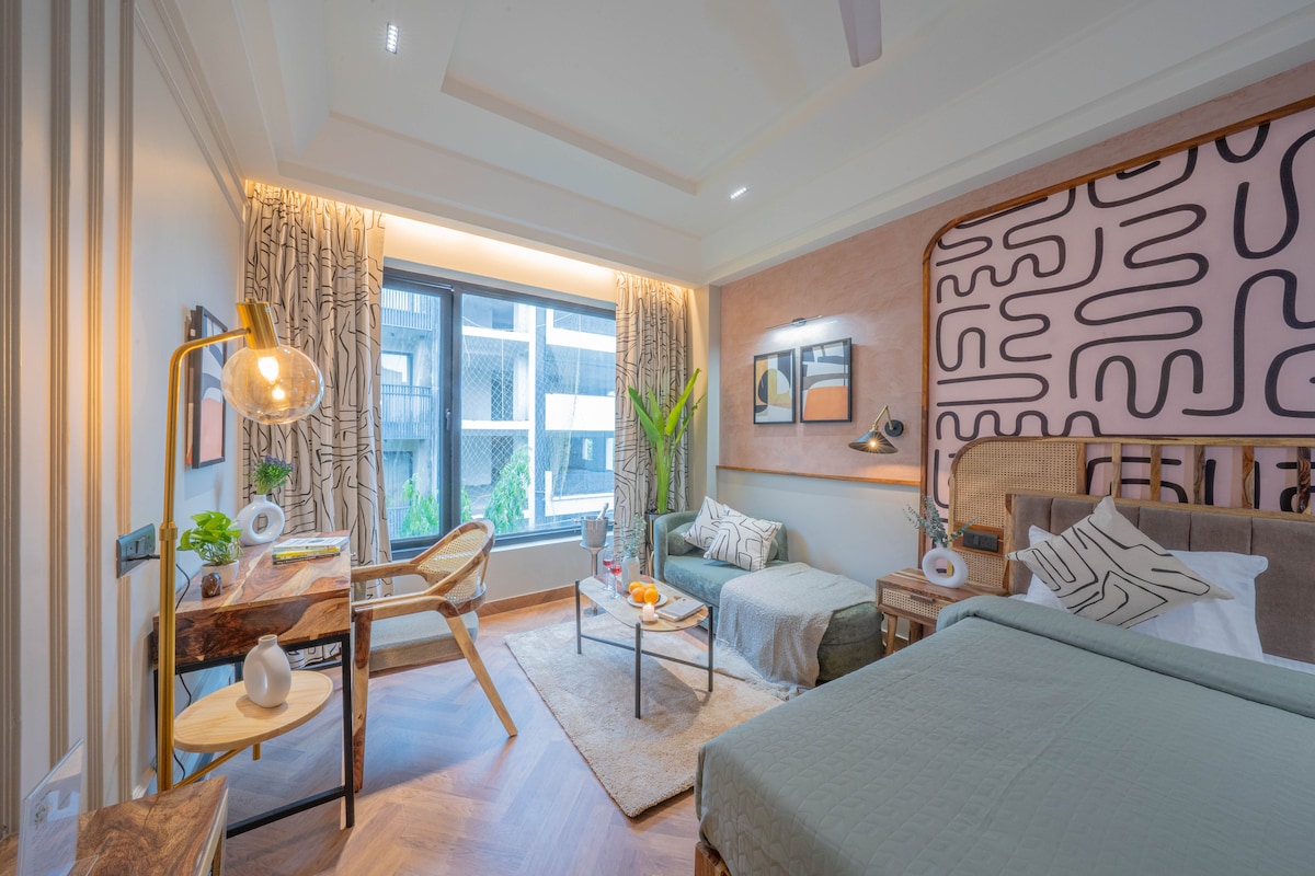 A well-furnished studio room features a spacious bed and a cozy sofa beside a center table. Natural light flows through a large window. Decorative wall art adds character, while a wooden desk provides a work area. A plant brings a touch of nature to the space.