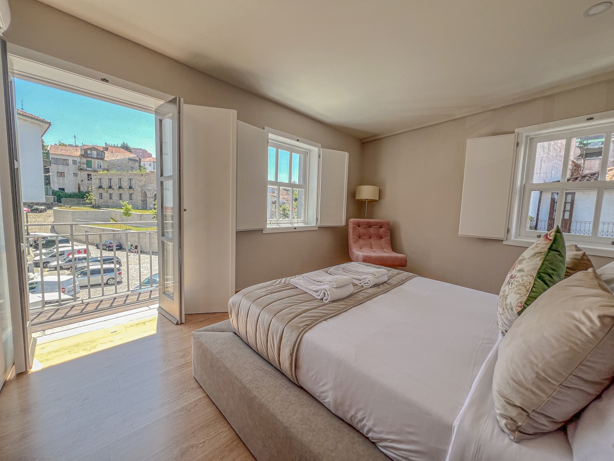 A well-lit bedroom features a spacious bed with neutral-toned linens. Light streams in from two open windows, enhancing the room's airy feel. A pink accent chair is positioned beside the door leading to a balcony, which offers views of the surrounding area.
