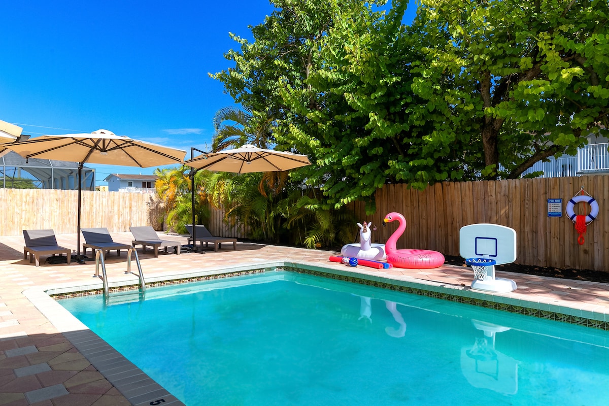 A private backyard is featured, showcasing a clear blue pool surrounded by lounge chairs. Two umbrellas provide shade, while pool floats, including a unicorn and a flamingo, add a playful touch. Lush greenery and a wooden fence create a serene environment.
