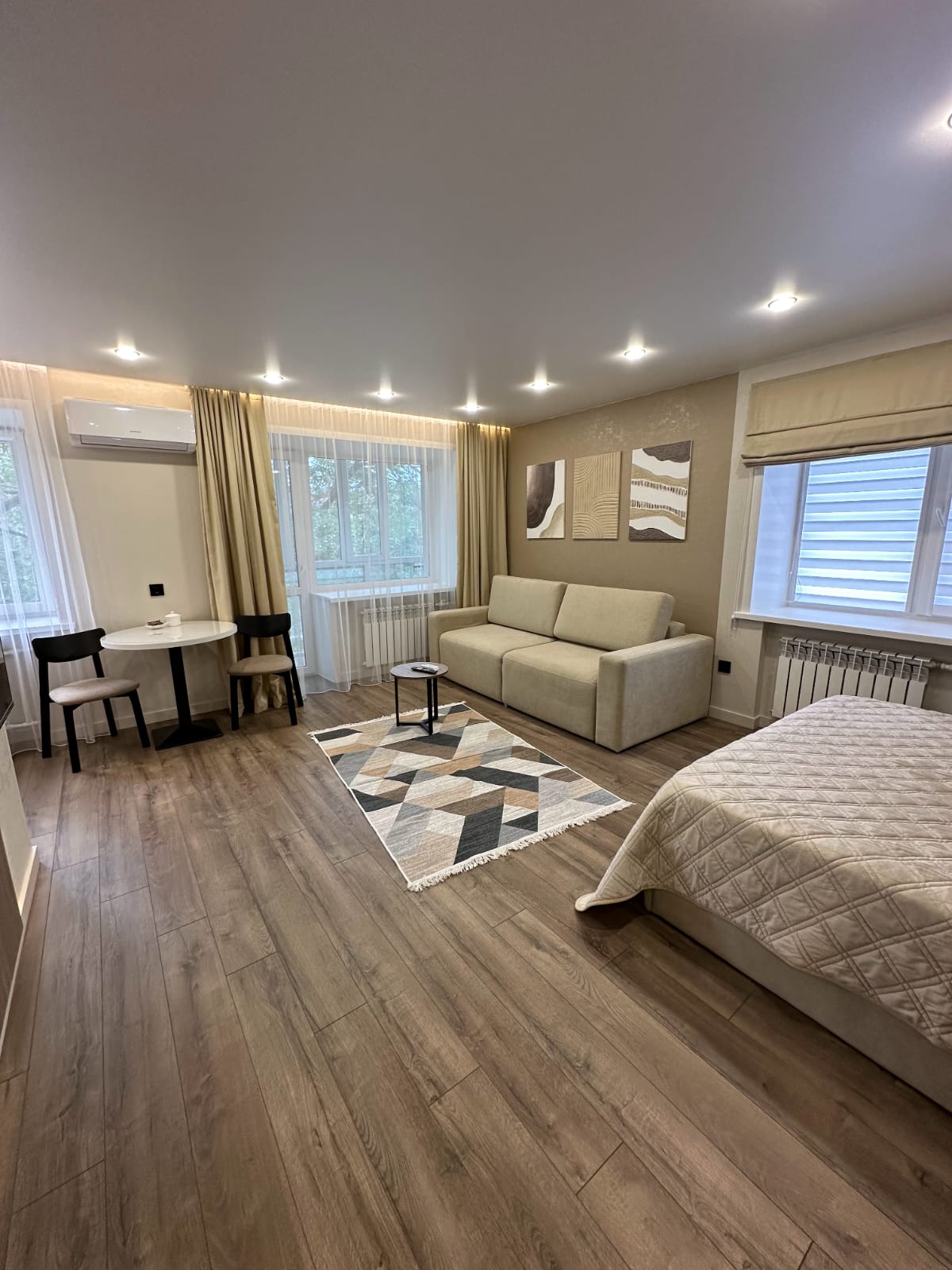 A compact living space is presented with a neutral-colored sofa and a small dining table. Natural light filters through two windows, illuminating the room's wooden floors and subtle wall art. A patterned rug adds texture, creating a cozy, functional environment.