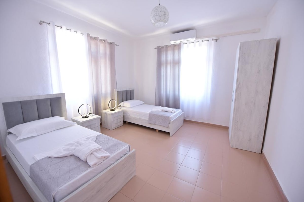 A bright bedroom features two spacious beds, one of which is positioned against a window adorned with flowing curtains. Soft, neutral-toned walls complement the minimalist design. A light fixture hangs from the ceiling, and a wardrobe stands against one wall.