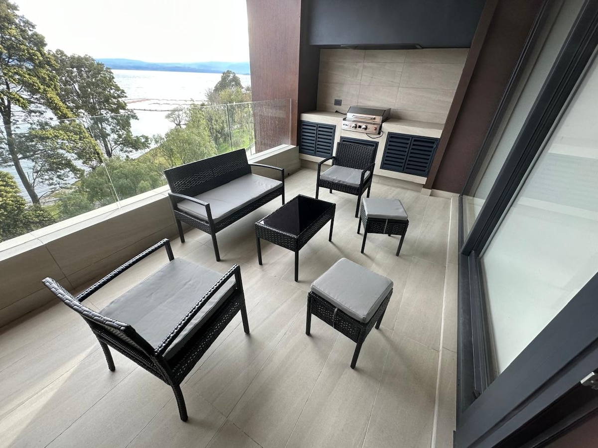 An outdoor seating area features a set of black wicker furniture, including a spacious couch, chairs, and a table. A grill is positioned nearby, and the area is surrounded by glass panels, offering views of the lake and greenery in the distance.