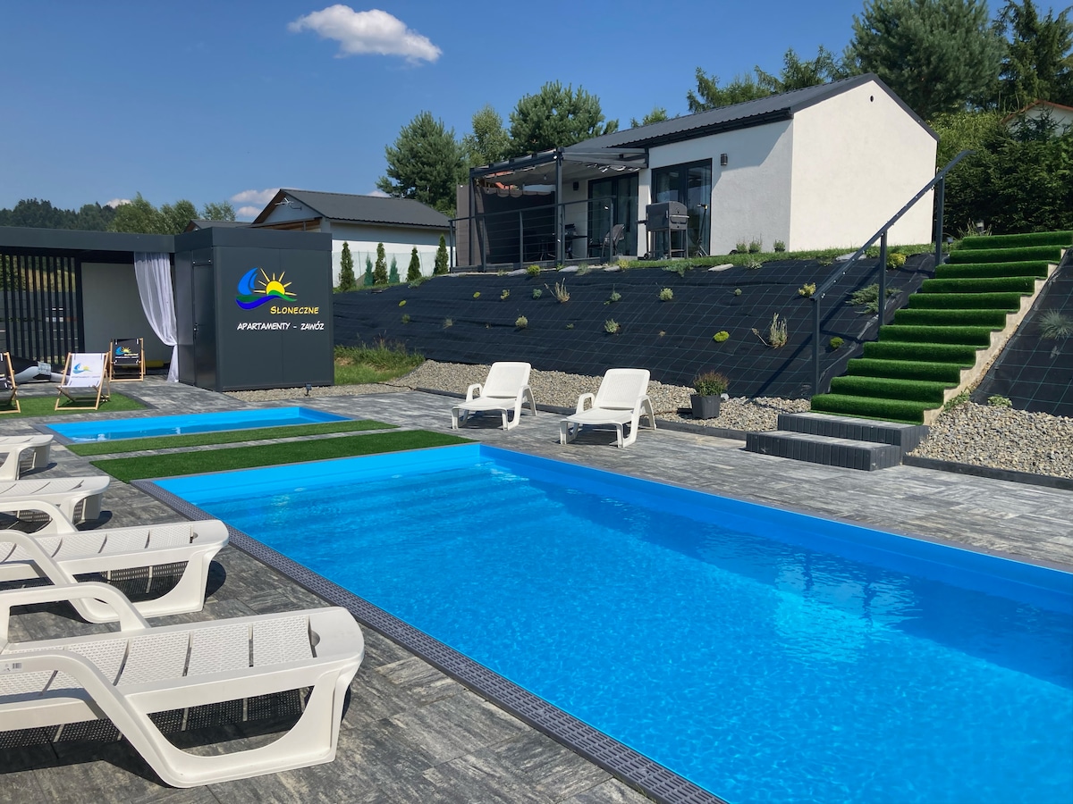A serene outdoor space features two inviting swimming pools surrounded by a tiled patio. Lounge chairs are positioned near the pools, and a landscaped area includes stairs leading to a contemporary building with a terrace. Soft greenery complements the setting under a clear blue sky.