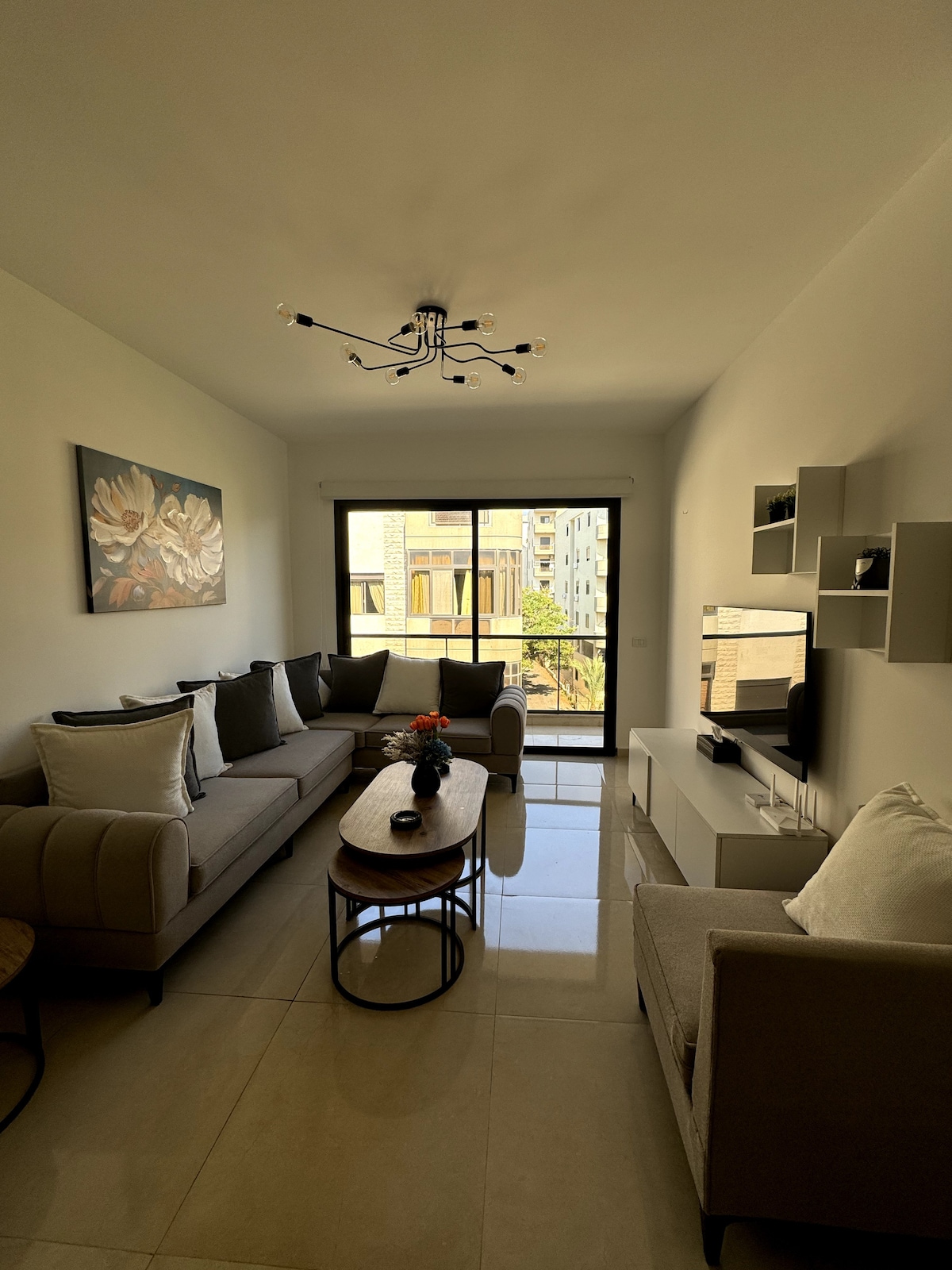 A modern living area features a comfortable sectional sofa alongside a chair, with a coffee table at the center. Large windows provide natural light and a view of the surroundings. A flat-screen TV is mounted on the wall alongside minimalist shelving.
