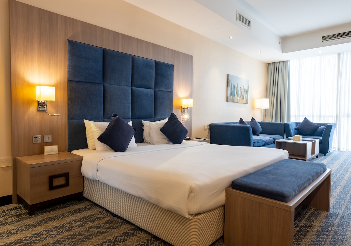 A spacious room featuring a king bed with a padded headboard, complemented by accent pillows. A cozy sitting area includes a sofa and armchair, providing comfort. Natural light filters in through large windows, while a modern bedside table and stylish bench complete the relaxing atmosphere.