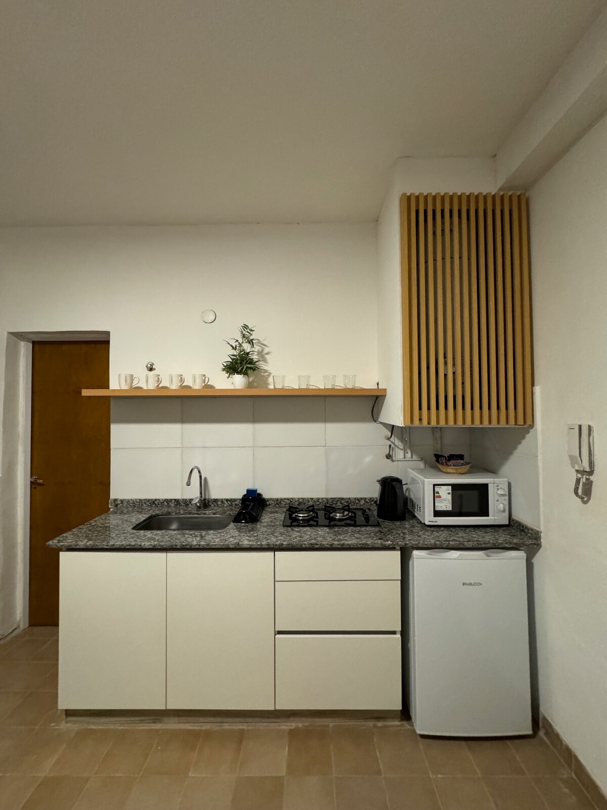 A compact kitchen area features a countertop with a sink, a gas stove, and a microwave. Storage is provided by upper cabinets, while a small refrigerator sits discreetly below the counter. Decorating items are placed on the shelf above, contributing to the neat arrangement.