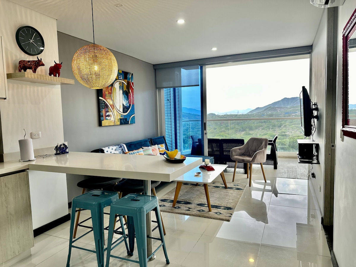 A modern living space features a light-filled area with an open layout. A spacious sofa in blue is positioned near large glass doors, revealing scenic mountain views. A minimalist dining table with turquoise stools complements the contemporary decor, accented by artwork and a stylish pendant light.
