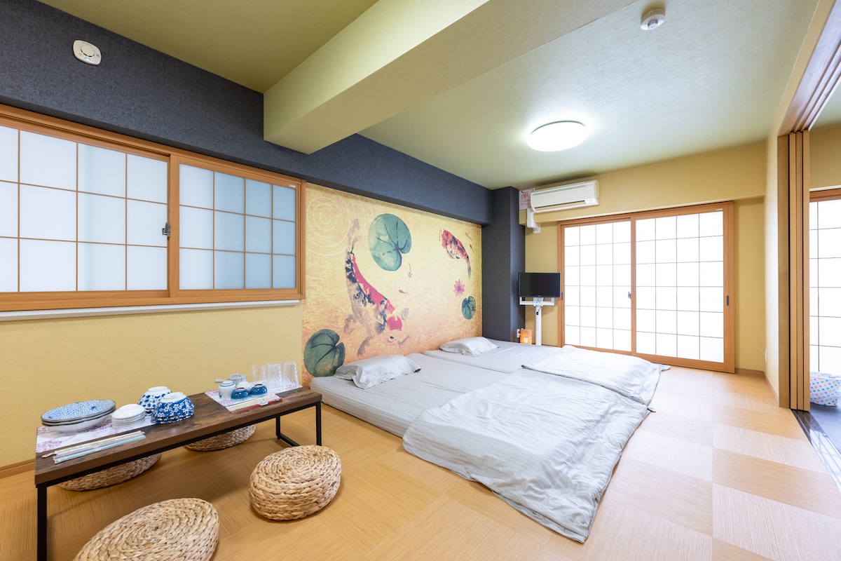 The spacious room features a minimalist design with tatami flooring and sliding shoji doors. Soft bedding is arranged on the floor, complemented by a decorative wall with floral art. A low table is set with dishware, and a television is mounted nearby. Natural light streams in through the windows.