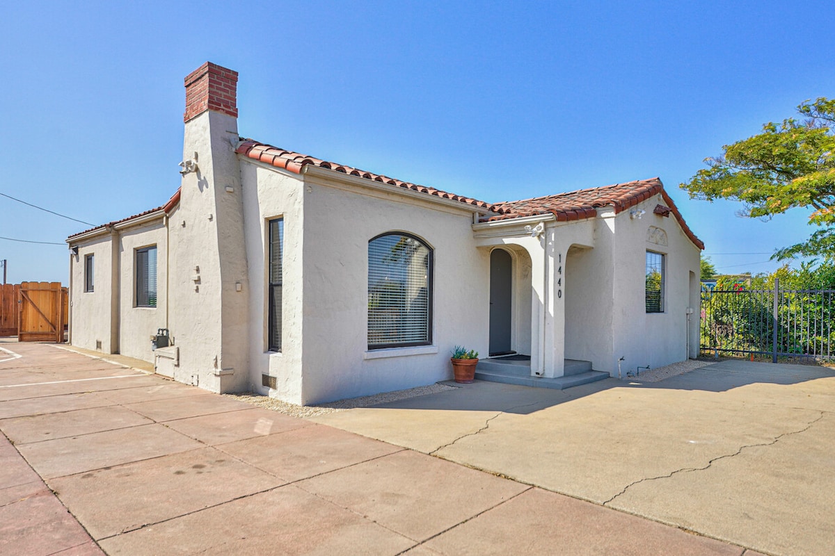 A Spanish-style home exterior is shown, featuring a light stucco facade and a red-tiled roof. A welcoming entryway is framed by arched windows, while a small potted plant adds a touch of greenery. A spacious driveway accommodates parking, complemented by a fenced area in the background.
