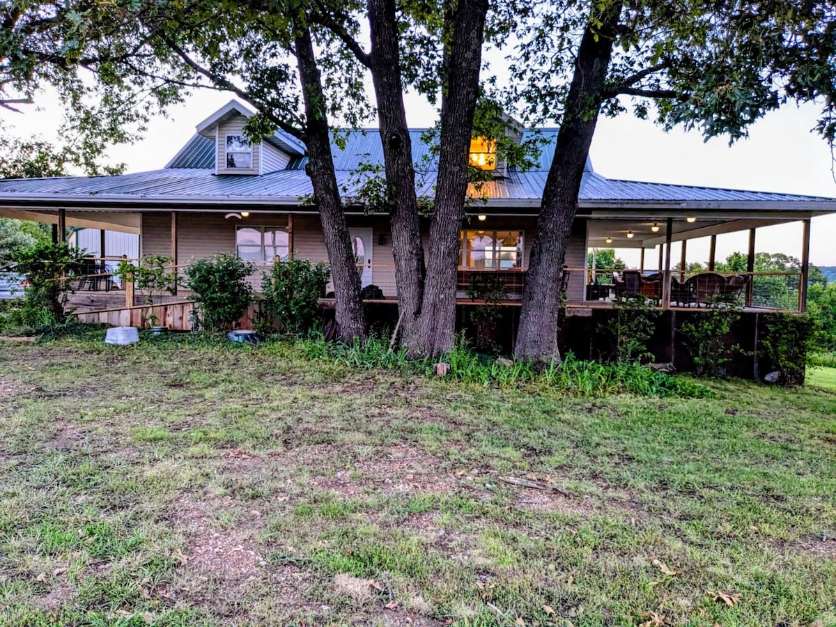 The farmhouse is surrounded by lush greenery, featuring a spacious wraparound porch supported by two large trees. Soft evening light glows from the windows, highlighting the inviting structure, which includes areas for outdoor seating. The yard is kept natural with a well-maintained grassy area.