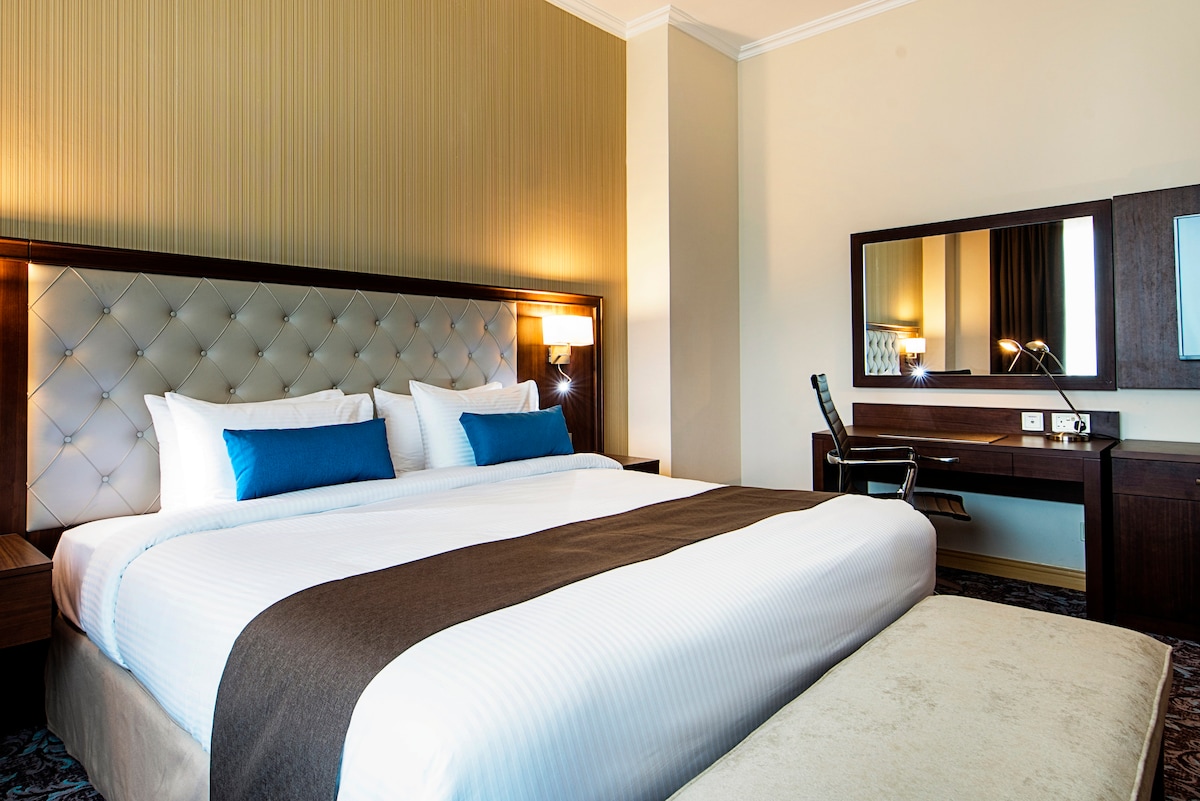 A spacious executive room features a large king bed with decorative pillows and elegant headboard. A comfortable sitting area with a plush sofa is visible. A functional desk with a swivel chair is positioned near a mirror, providing a practical workspace.
