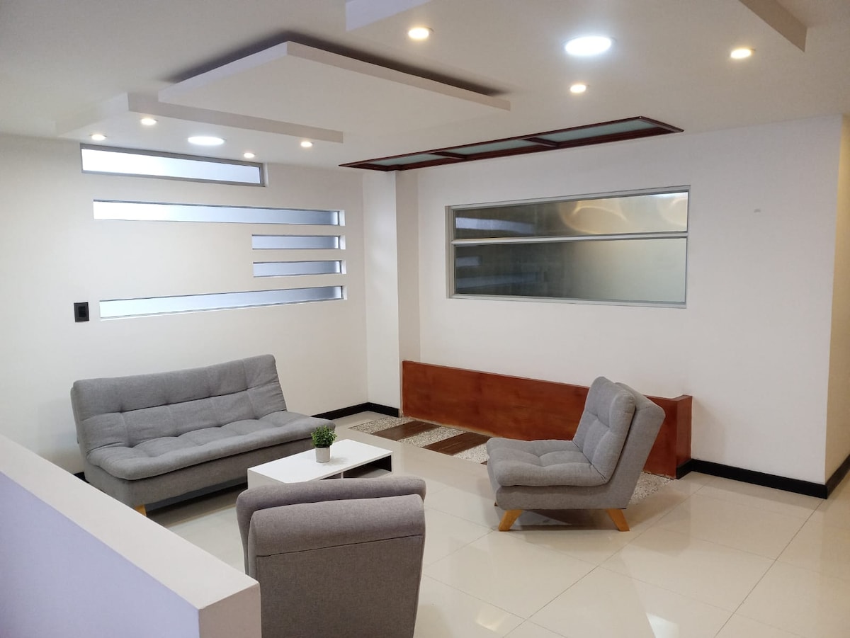 A modern living area features two gray sofas and a small white coffee table. Natural light floods the space through large windows, complemented by recessed lighting on the ceiling. A decorative rug is positioned on the polished floor, adding texture and warmth.