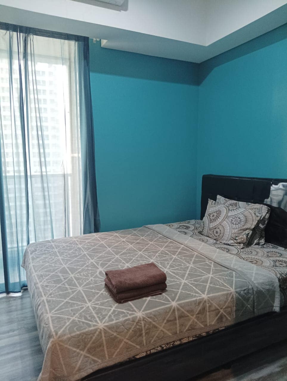 A serene bedroom features a spacious bed dressed in neutral-toned bedding and a decorative pillow. A rolled towel rests at the foot of the bed. Light filters through sheer curtains, which frame a glass door leading to a balcony with a city view.