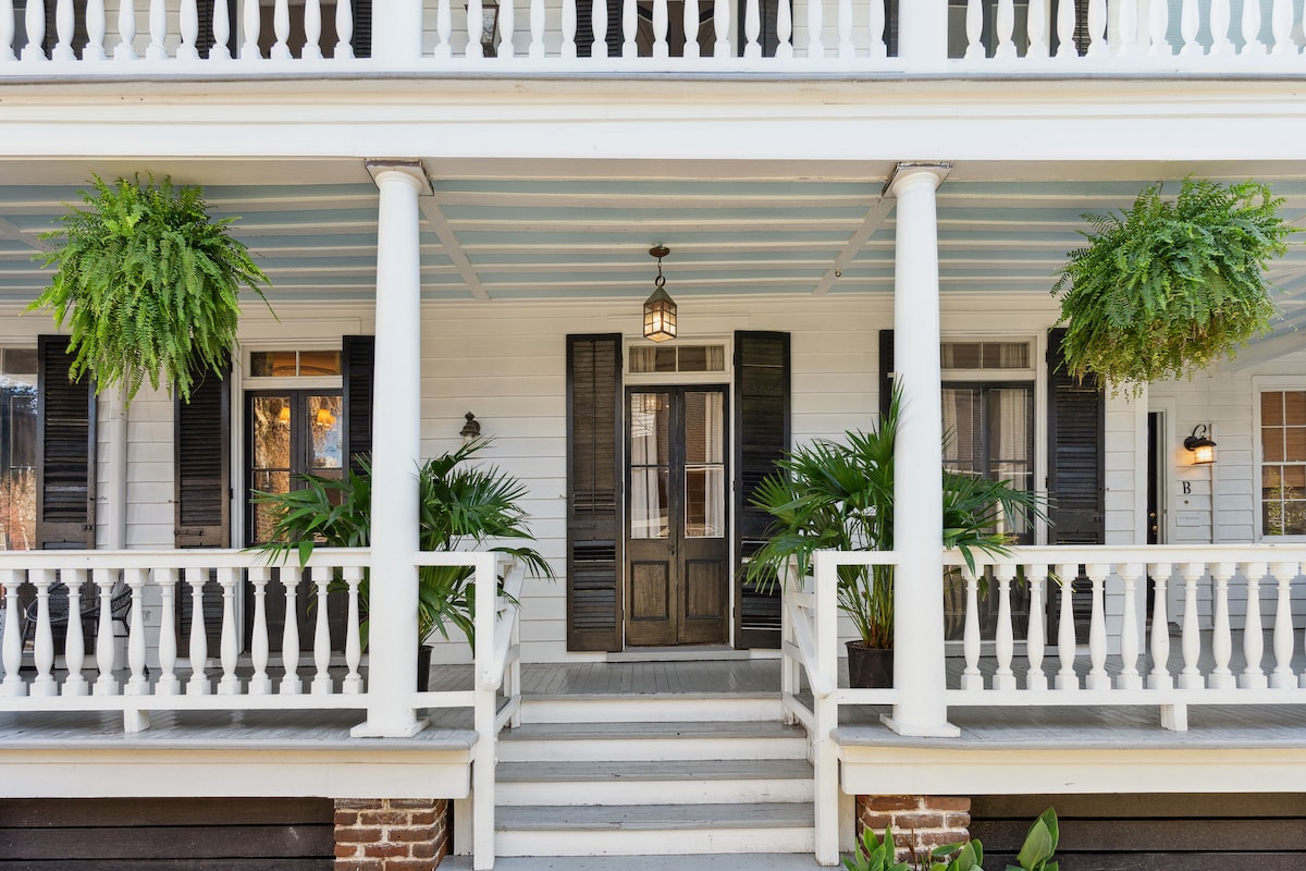 The entrance showcases a welcoming porch with large wooden doors framed by black shutters. Lush ferns hang from white columns, complementing the greenery on either side of the steps. The porch features elegant railings and a hanging lantern, enhancing the historic charm of the property.