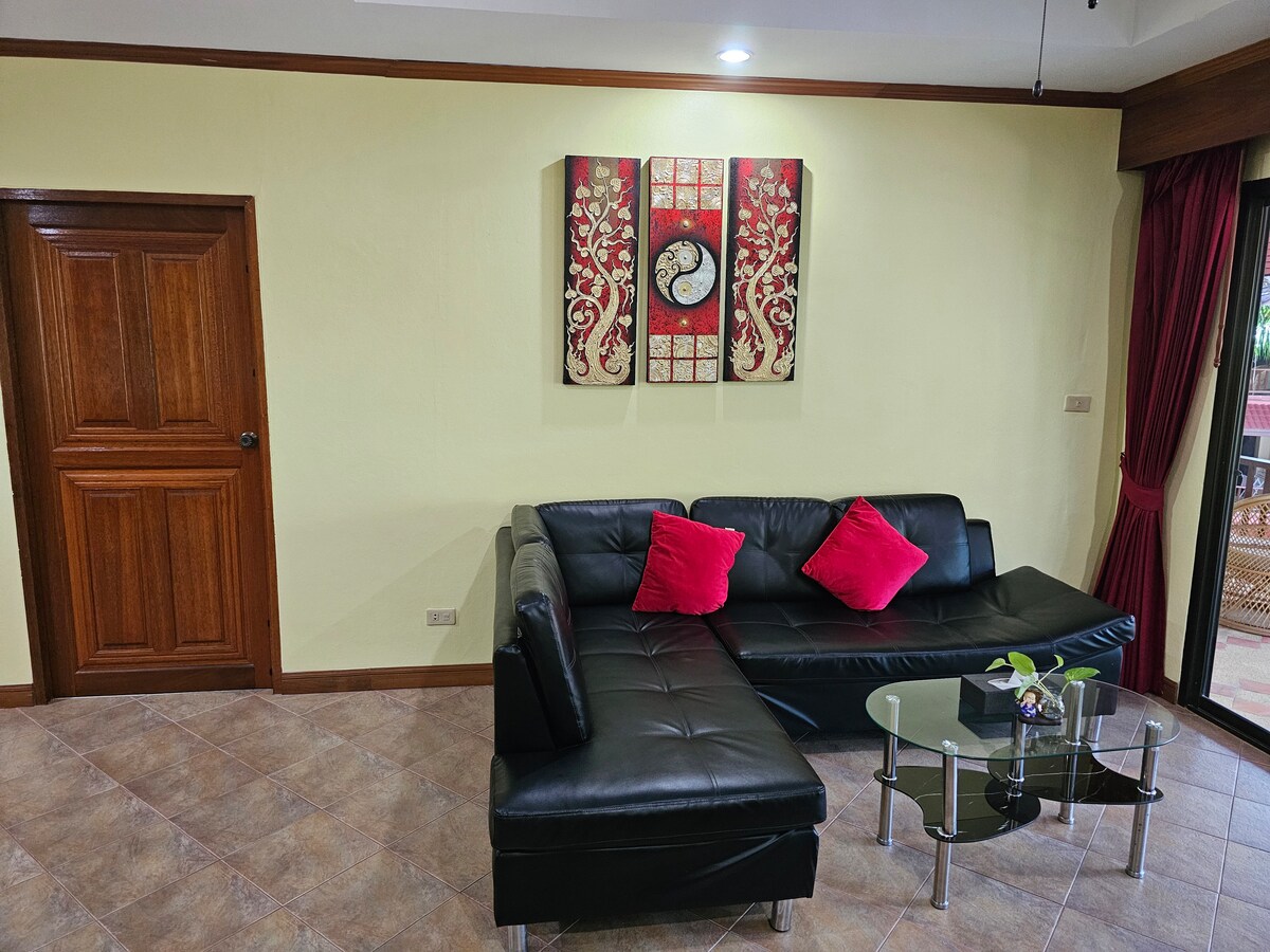 A lounge area features a dark leather sectional sofa adorned with red accent pillows. A small glass coffee table is positioned in front, and a decorative wall art piece enhances the atmosphere. Soft light is emitted from overhead fixtures, illuminating the room's warm colors.