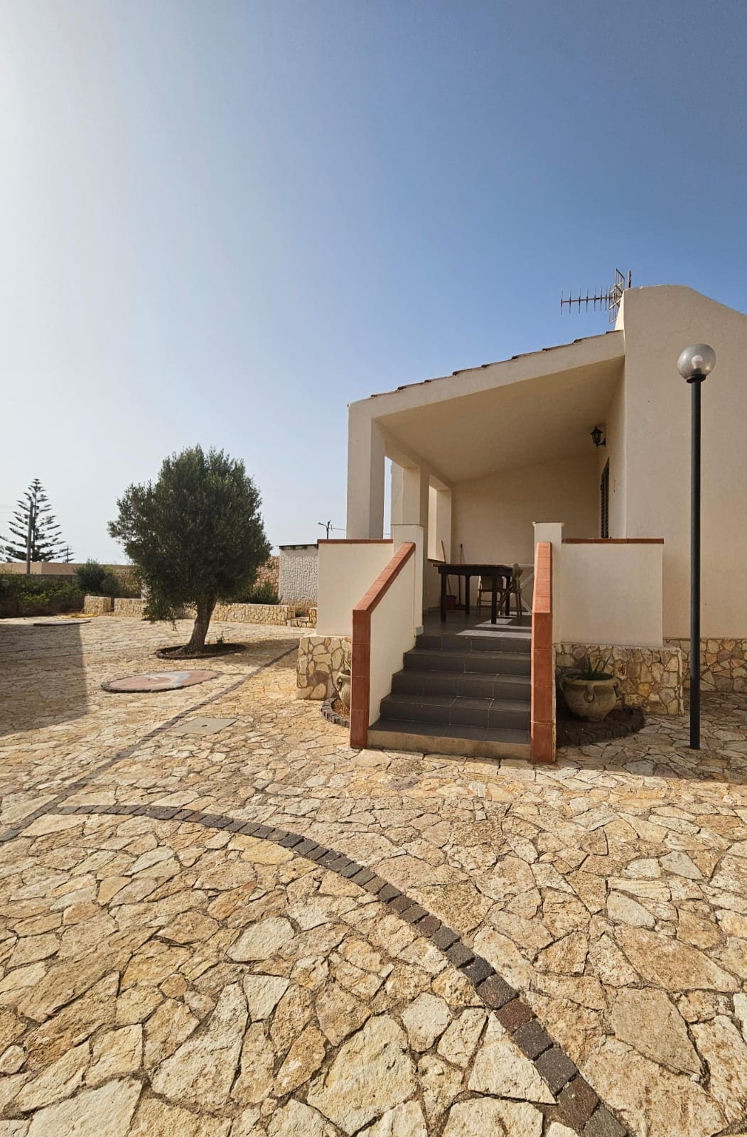 A spacious exterior area features a stone pathway leading to a covered terrace with a table and chairs. A potted plant is positioned beside the steps, while a street lamp provides lighting. Surrounding greenery adds to the tranquil environment.