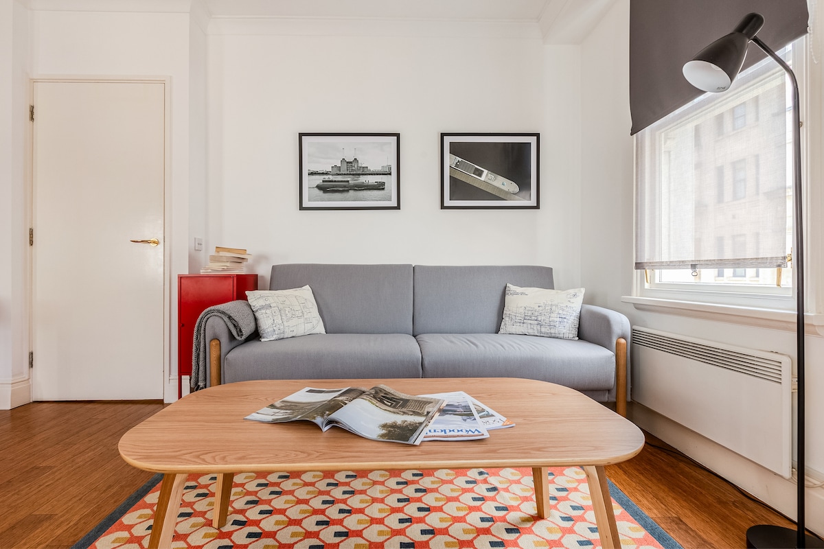A cozy living area features a light grey sofa adorned with cushions, positioned opposite a low wooden coffee table. On the wall, two framed artworks are displayed. Natural light streams in through a window covered by a roll-up shade, highlighting the warm wooden floor.