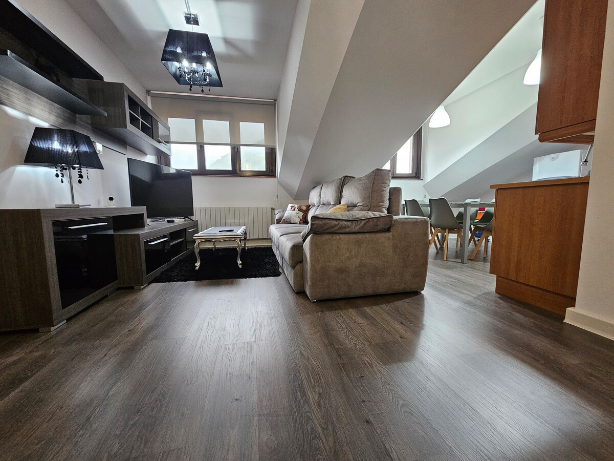 A modern living space features a comfortable sofa facing a wall-mounted television. Large windows allow natural light to brighten the room, while a sleek dining area is visible in the background. Wooden flooring adds warmth to the overall ambiance.