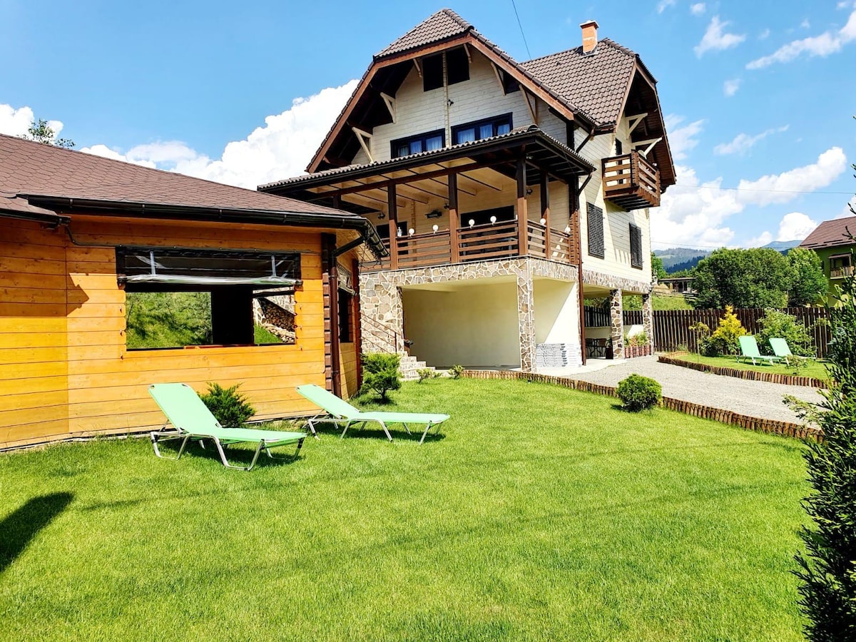 The exterior of the house showcases a two-story structure with a combination of wooden and stone elements. A spacious lawn features green loungers, while the wraparound porch offers seating, blending harmoniously with the surrounding landscape of trees and distant mountains.