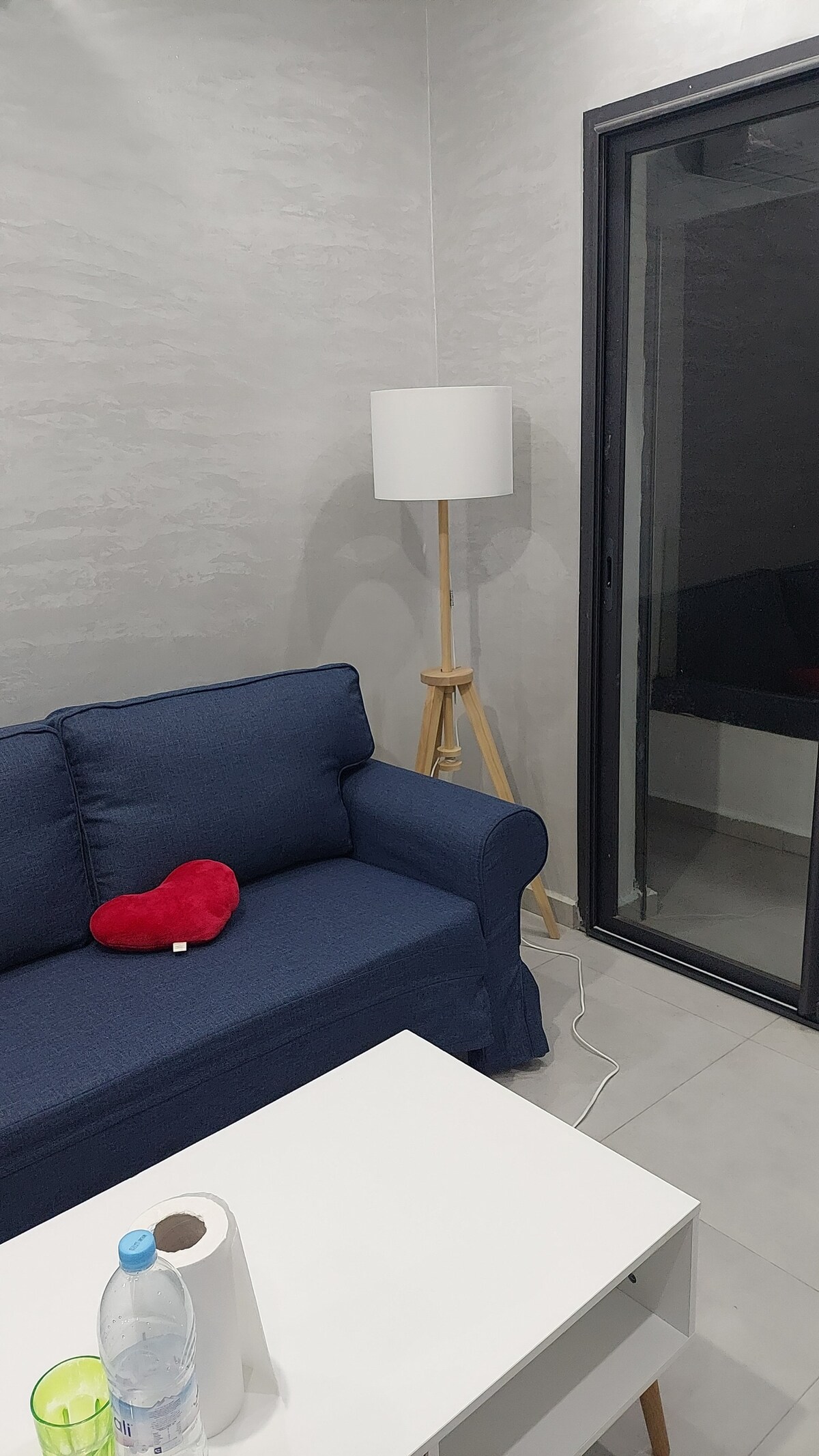 A cozy sitting area features a navy blue couch adorned with a heart-shaped cushion. A minimalist white lamp stands beside the couch on a wooden tripod. A small white coffee table holds a bottle of water and paper towels. A glass door leads to an outdoor space.